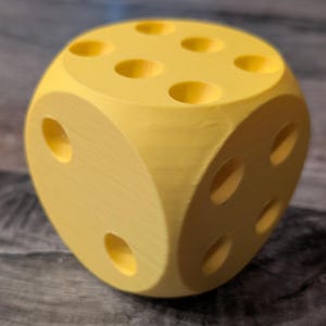 May include: A large, yellow die with rounded edges and recessed circular markings. The die is a solid colour and appears to be made of plastic. The die has the typical six sides, each with a different number of recessed circles.