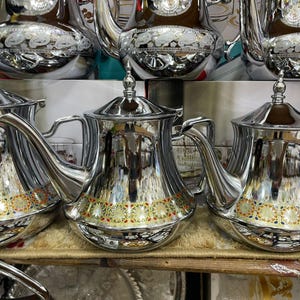 Peut inclure: Théières argentées brillantes avec des motifs décoratifs. Les théières ont un corps arrondi, des becs courbés et des poignées ornées. Une bande de motifs floraux colorés orne la base de chaque théière. Les théières sont exposées sur une étagère.