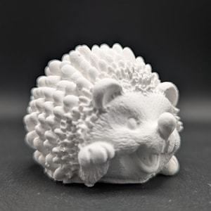 Könnte beinhalten: Eine weiße Keramik-Igel-Figur mit detaillierten Stacheln und einem lächelnden Gesicht. Die kleine Skulptur hat sichtbare Pfoten und Ohren, vor einem dunklen Hintergrund.