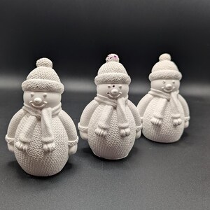Könnte beinhalten: Drei weiße Schneemannfiguren mit Strickmützen und Schals. Jeder Schneemann hat ein lächelndes Gesicht und Knopfaugen. Die Hüte haben einen kleinen, dekorativen Bommel oben. Die Schneemänner sind in einer Reihe vor einem dunklen Hintergrund angeordnet.