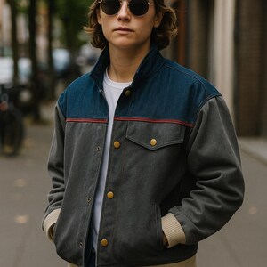 Puede incluir: Una persona con una chaqueta gris y azul con una raya roja en el pecho, una camiseta blanca y vaqueros azules. La chaqueta tiene botones dorados y ribete beige. También lleva una gorra de béisbol marrón y gafas de sol redondas.