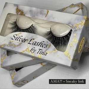 Può includere: Scatole di ciglia finte con un design marmorizzato e dorato. Le scatole sono etichettate "Silver Lashes by Tina" e contengono ciglia nere. Il testo "AMA!!! = Sneaky link" è in basso.