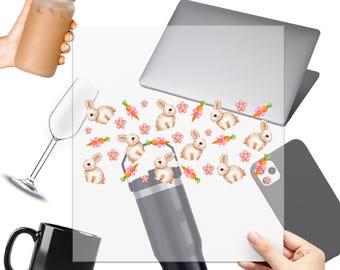 Op de afbeelding: Een heldere UV DTF-sticker met een herhalend patroon van witte konijntjes, wortels en roze bloemen. De sticker wordt getoond op een laptop, een beker, een koffiemok, een wijnglas, een telefoon en een glas met een rietje. De tekst "UV DTF STICKER" staat bovenaan.