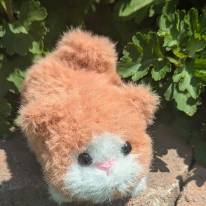 May include: A small, fuzzy plush toy shaped like a cat with orange fur, a white patch on its face, a small pink nose, and two round black eyes. The plush has a soft, textured appearance and is positioned in a low, crouching posture.