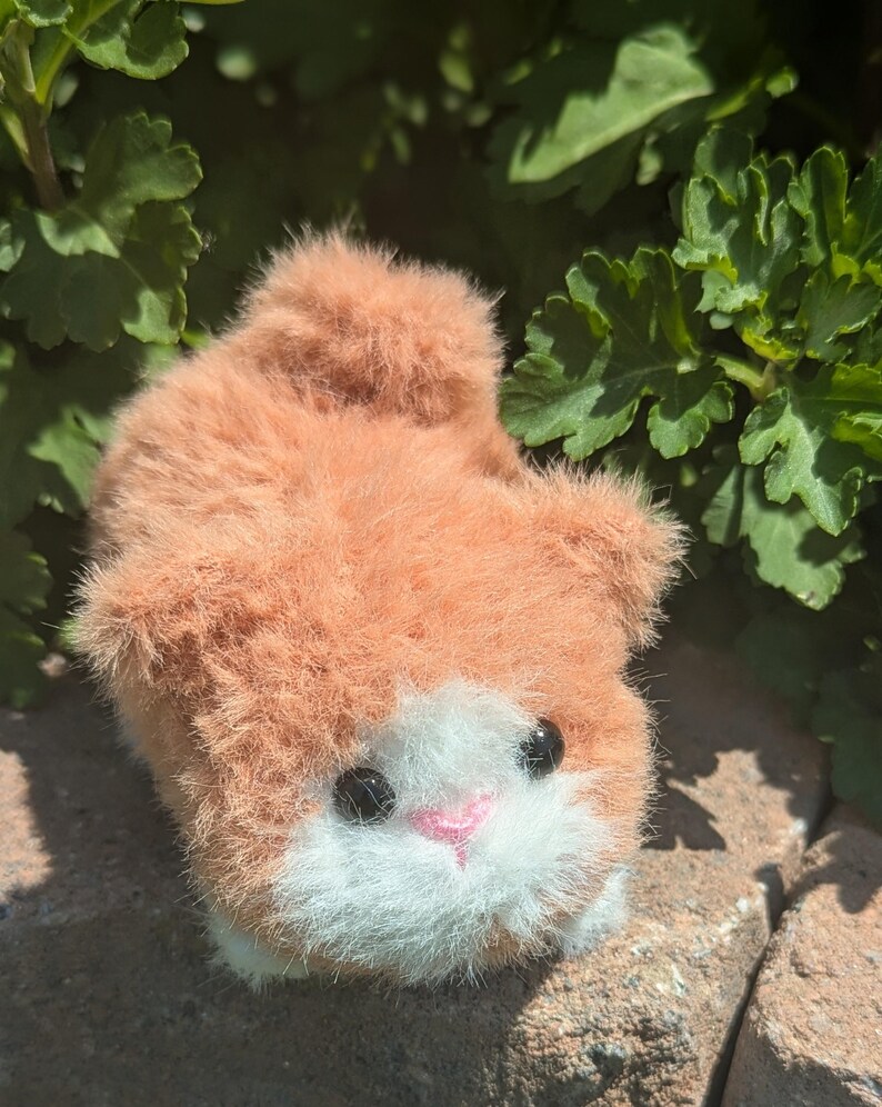 May include: A small, fuzzy plush toy shaped like a cat with orange fur, a white patch on its face, a small pink nose, and two round black eyes. The plush has a soft, textured appearance and is positioned in a low, crouching posture.