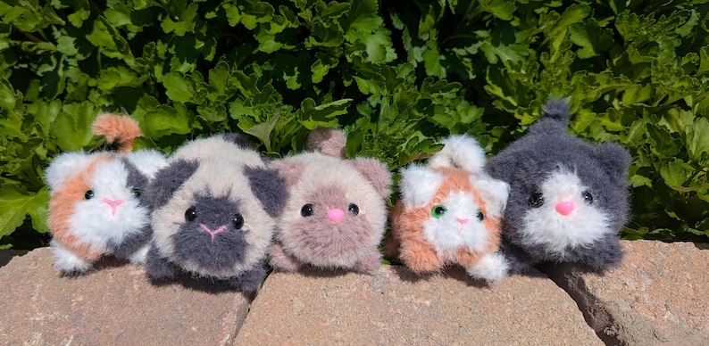 May include: Five small, fuzzy, handmade plush cat toys arranged in a row. The cats feature varied color patterns including calico, grey and white, light brown, orange and white, and solid dark grey, all with small pink noses and black eyes.