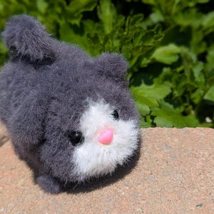 May include: A small, fuzzy, dark gray plush cat toy with a white face patch, a bright pink plastic nose, and two small black plastic eyes. The plush has a rounded body shape with a short, upright tail and a soft, textured, yarn-like appearance.