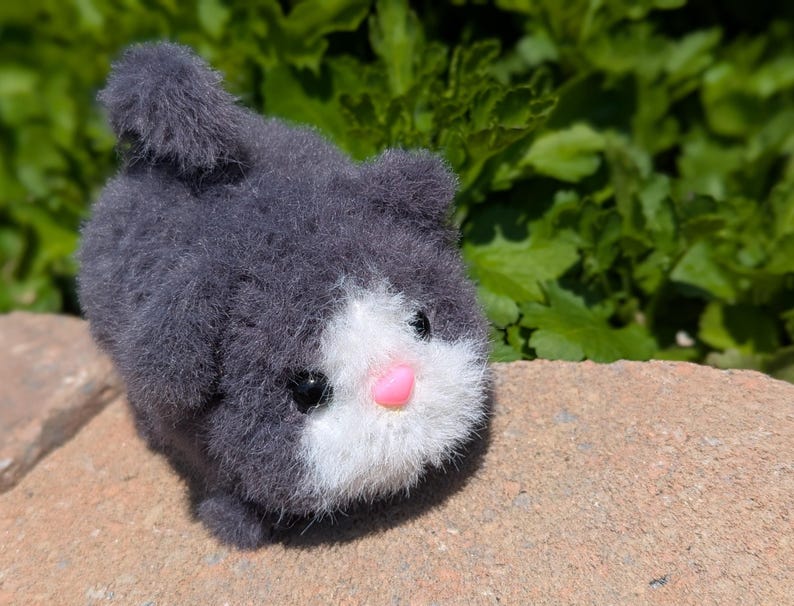 May include: A small, fuzzy, dark gray plush cat toy with a white face patch, a bright pink plastic nose, and two small black plastic eyes. The plush has a rounded body shape with a short, upright tail and a soft, textured, yarn-like appearance.