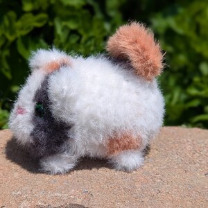 May include: A small, fuzzy, plush guinea pig figurine featuring a tri-color pattern of white, dark gray, and light orange fur. The creature has a small pink nose, dark green eyes, and a rounded, textured body with a prominent orange tail tuft.