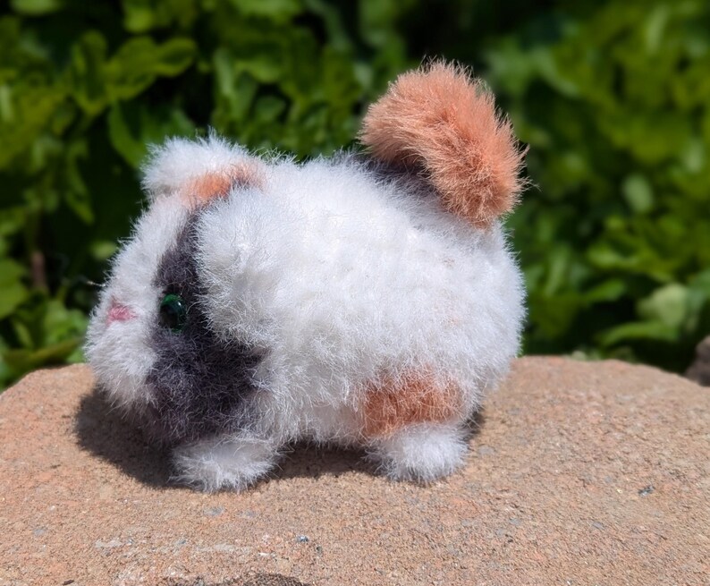 May include: A small, fuzzy, plush guinea pig figurine featuring a tri-color pattern of white, dark gray, and light orange fur. The creature has a small pink nose, dark green eyes, and a rounded, textured body with a prominent orange tail tuft.