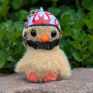 May include: A plush yellow chick keychain wearing a white helmet with red flames and a black face mask. The chick has black eyes, an orange beak, and orange feet. The keychain is made of soft, fuzzy material.