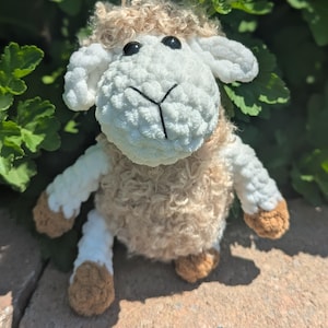 May include: A plush sheep toy crafted from textured yarn, featuring a cream-colored curly body, a white face with black plastic eyes, and a stitched black nose. The limbs are white with brown-colored feet, and the head has floppy white ears.
