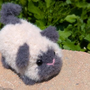 May include: A small, fuzzy, handcrafted plush animal figure with a cream-colored body and dark gray accents on the ears, face, tail, and feet. It features two round black eyes and a small, stitched pink nose, resembling a miniature cat or kitten.