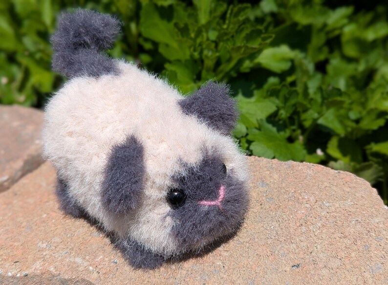 May include: A small, fuzzy, handcrafted plush animal figure with a cream-colored body and dark gray accents on the ears, face, tail, and feet. It features two round black eyes and a small, stitched pink nose, resembling a miniature cat or kitten.