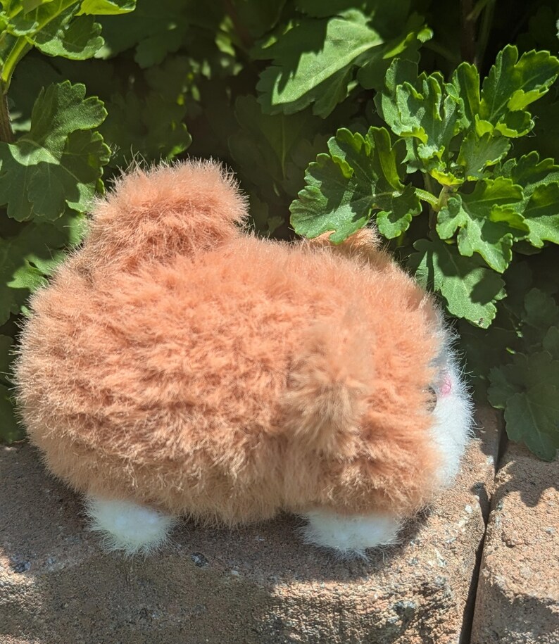 May include: A small, round, plush toy shaped like a fluffy animal with light brown, shaggy fur, a prominent tail, and small white paws. The toy features a white face area with a small pink nose and is positioned on a textured stone surface.