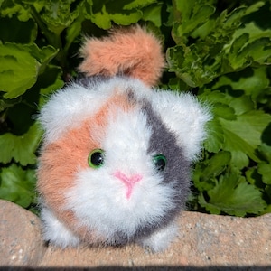 May include: A small, round, plush toy shaped like a calico cat with fluffy white, orange, and dark gray fur. The toy features bright green plastic eyes and a small, stitched pink nose, with a tuft of orange fur on top of its head.