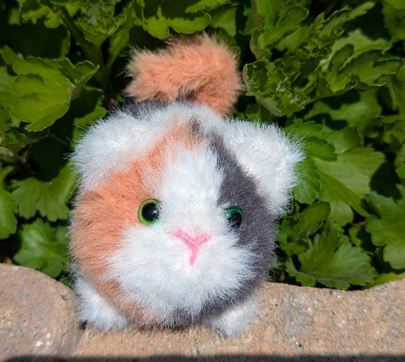 May include: A small, round, plush toy shaped like a calico cat with fluffy white, orange, and dark gray fur. The toy features bright green plastic eyes and a small, stitched pink nose, with a tuft of orange fur on top of its head.