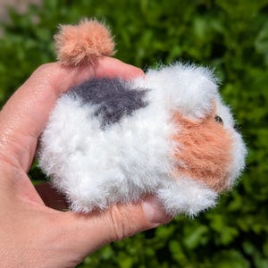 May include: A small, fuzzy, handcrafted pom-pom animal figure resembling a guinea pig. The creature features a white base with patches of dark gray and light orange fur, a small orange pom-pom tail, and a visible dark eye on the side of its head.