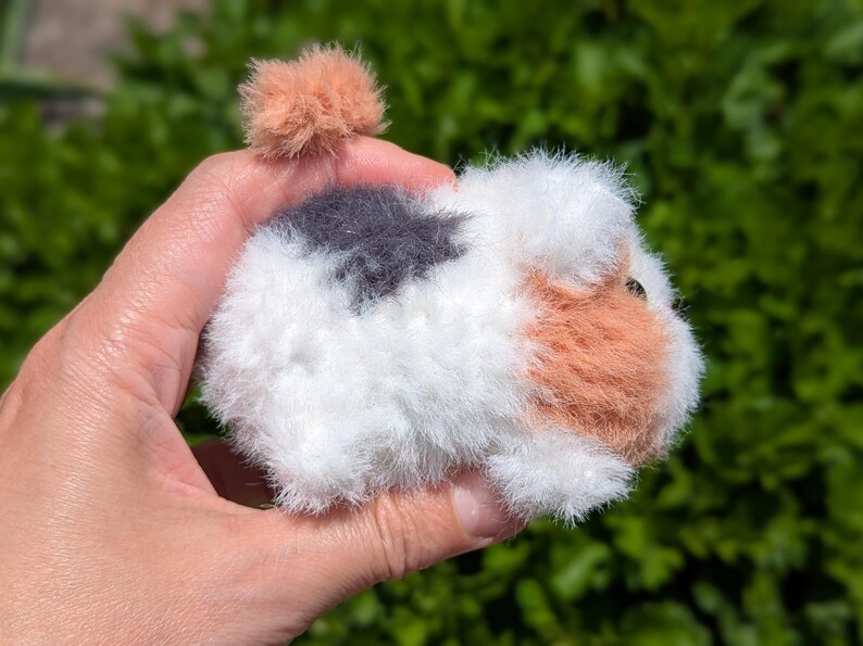 May include: A small, fuzzy, handcrafted pom-pom animal figure resembling a guinea pig. The creature features a white base with patches of dark gray and light orange fur, a small orange pom-pom tail, and a visible dark eye on the side of its head.