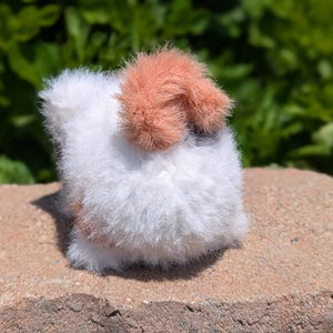 May include: A small, round, plush animal figure crafted from fuzzy white yarn with a prominent patch of soft, peach-colored fur on the top. The texture is highly fibrous and dense, creating a rounded, compact shape that sits firmly on a flat surface.