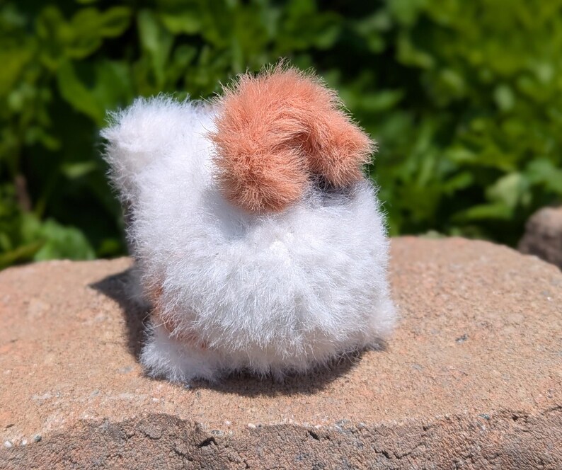 May include: A small, round, plush animal figure crafted from fuzzy white yarn with a prominent patch of soft, peach-colored fur on the top. The texture is highly fibrous and dense, creating a rounded, compact shape that sits firmly on a flat surface.