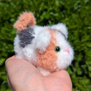May include: A small, fluffy, handcrafted pom-pom animal figure featuring a calico pattern of white, orange, and grey synthetic fibers. The creature has a small, pink stitched nose and bright green plastic eyes, with a short, upright tail.