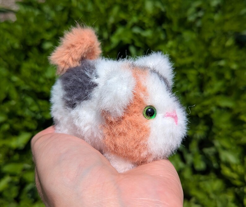 May include: A small, fluffy, handcrafted pom-pom animal figure featuring a calico pattern of white, orange, and grey synthetic fibers. The creature has a small, pink stitched nose and bright green plastic eyes, with a short, upright tail.