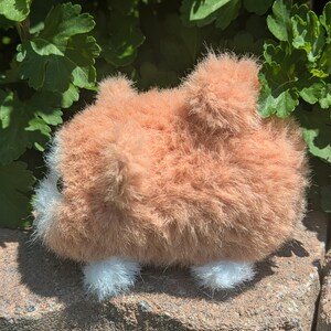 May include: A small, fuzzy plush toy shaped like a hamster with light orange fur, a white face patch, and white paws. The toy has a rounded body, a small tail, and a visible dark eye, presented in a side profile view with a soft, textured appearance.