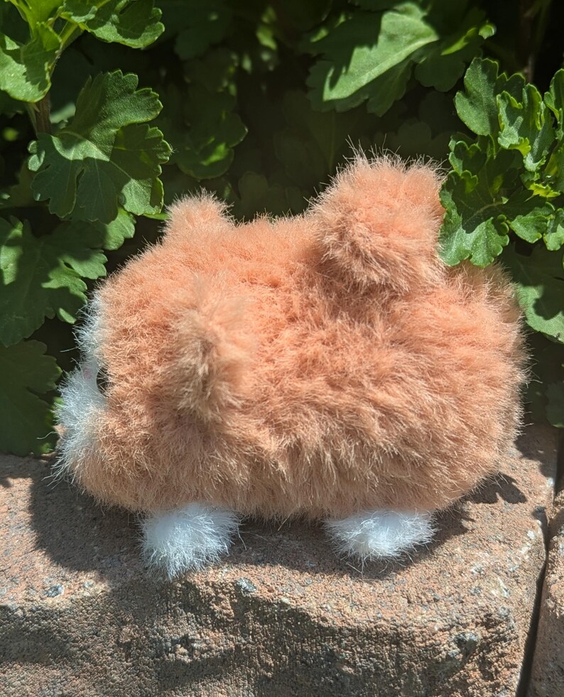 May include: A small, fuzzy plush toy shaped like a hamster with light orange fur, a white face patch, and white paws. The toy has a rounded body, a small tail, and a visible dark eye, presented in a side profile view with a soft, textured appearance.