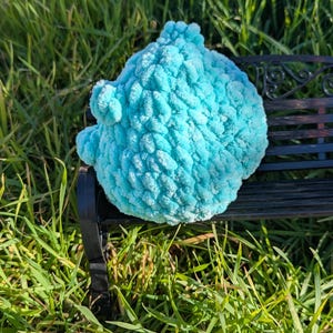 May include: A plush, light blue, cloud-shaped decorative item rests on a small, black, wrought-iron bench. The item has a textured, bumpy surface. The bench is set in a bed of green grass.