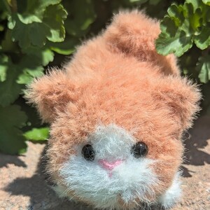 May include: A small, fuzzy, plush animal figurine with orange and white fur. The creature features two round, black, glossy eyes and a small, triangular, pink stitched nose. The texture of the material is soft and long-haired, giving it a rounded, compact appearance.