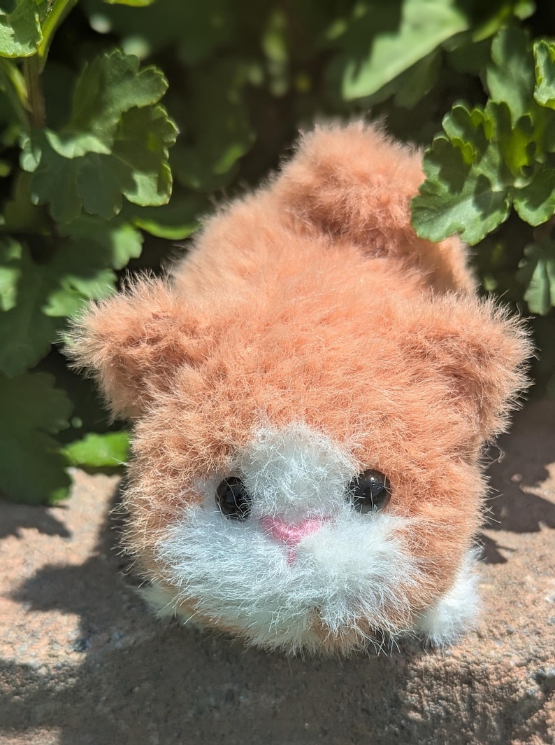 May include: A small, fuzzy, plush animal figurine with orange and white fur. The creature features two round, black, glossy eyes and a small, triangular, pink stitched nose. The texture of the material is soft and long-haired, giving it a rounded, compact appearance.