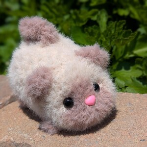 May include: A small, fuzzy, cream-colored plush animal with soft, dusty-rose accents on its ears, tail, and face. It features two round, black plastic eyes and a small, smooth, bright pink heart-shaped nose, giving it a rounded, compact appearance.