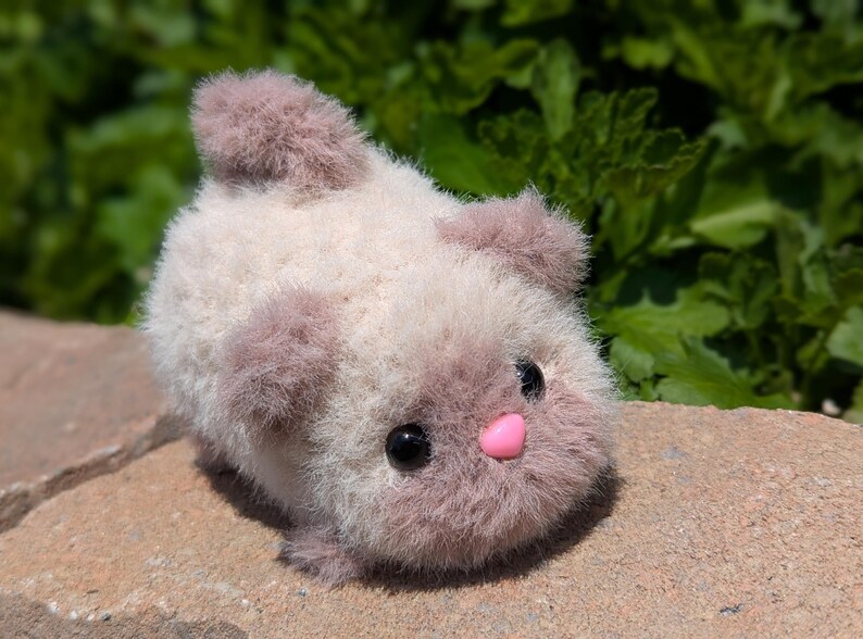 May include: A small, fuzzy, cream-colored plush animal with soft, dusty-rose accents on its ears, tail, and face. It features two round, black plastic eyes and a small, smooth, bright pink heart-shaped nose, giving it a rounded, compact appearance.