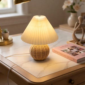 Peut inclure: Une petite lampe de table décorative avec une base en rotin tressé et un abat-jour plissé blanc cassé. La lampe est posée sur un bureau avec une surface imitation marbre, à côté d'un livre intitulé "In Paris".