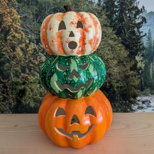 Könnte beinhalten: Ein Stapel aus drei Keramik-Halloween-Kürbissen in Orange, Grün und Creme. Jeder Kürbis hat ein geschnitztes Gesicht mit dreieckigen Augen und einem lächelnden Mund. Der obere Kürbis hat orangefarbene Streifen, der mittlere ist grün und der untere ist orange.