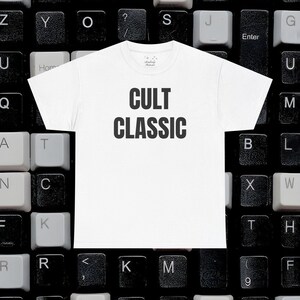 May include: White crew neck t-shirt with the words "CULT CLASSIC" printed in bold black letters. The shirt is displayed on a background of black and white computer keyboard keys.
