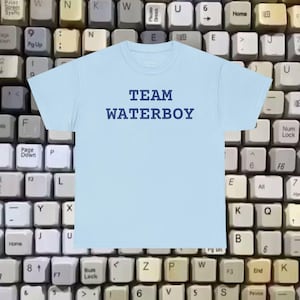 May include: A pale blue t-shirt featuring the navy blue text "TEAM WATERBOY". The shirt is set against a backdrop of old-fashioned computer keyboard keys.