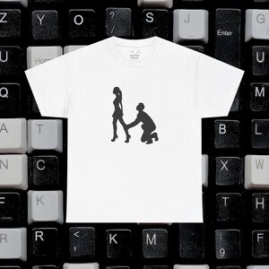 Puede incluir: Camiseta blanca con una silueta negra de una persona arrodillada que sostiene la mano de otra persona que se aleja. La camiseta se muestra sobre un fondo de teclado.