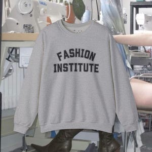 Puede incluir: Sudadera gris claro con la inscripción "FASHION INSTITUTE" en negro. La parte superior de cuello redondo y manga larga está hecha de un material suave.