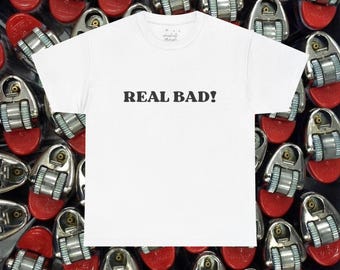 May include: A white short-sleeved t-shirt featuring the text "REAL BAD!" printed in black capital letters across the center of the chest. The shirt has a crew neckline and is displayed against a background of multiple metallic lighters.