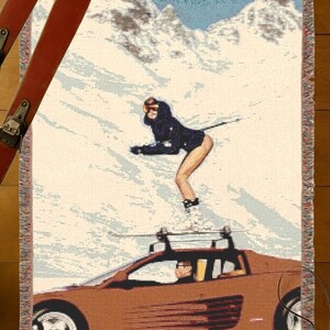 May include: Woven throw blanket depicting a skier in a black outfit on a snowy mountain, with a brown sports car below. Includes skis, goggles, and a camera. The blanket has a fringed edge.