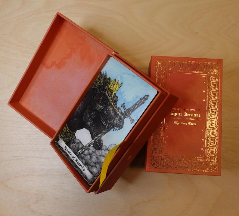May include: A tarot card with a dinosaur wearing a crown and holding a sword. The card is called "Queen of Swords" and is from the "Ignis Arcanae: The Fire Tarot" deck.