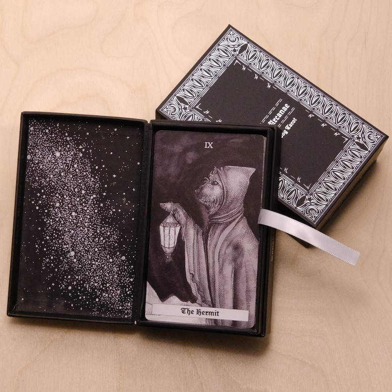 May include: A black tarot card deck box with a black and white illustration of a hooded figure holding a lantern. The card is labeled "The Hermit" and "IX".
