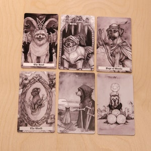 May include: A set of ten tarot cards featuring black and white illustrations of dogs. The cards are numbered with Roman numerals and have titles such as "The Devil", "Page of Wands", and "The World".