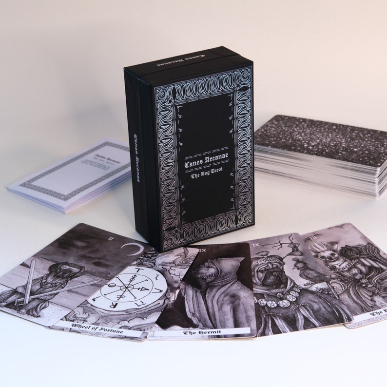 May include: A black box with silver lettering that reads "Canes Arcanae The Dog Tarot". The box is open and shows a deck of tarot cards with black and white illustrations of dogs.