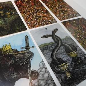 May include: A set of tarot cards with colorful illustrations. The cards feature a variety of imagery, including a dinosaur, a sword, and snakes. The cards are printed on thick cardstock and have a glossy finish.