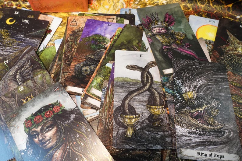 May include: A tarot card deck with a variety of cards spread out, including the King of Cups, the Two of Cups, and the Nine of Swords. The cards feature detailed illustrations with a dark and mystical aesthetic.