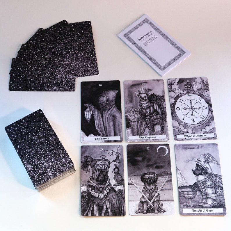 May include: A set of tarot cards with black and white illustrations of animals and figures. The cards feature the Hermit, the Empress, the Wheel of Fortune, the Knight of Cups, and other cards. The cards are laid out on a white surface.