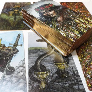 May include: A set of tarot cards with colorful illustrations. The cards feature a variety of images, including a dinosaur, a woman with flowers in her hair, and a snake with a cup. The cards are arranged in a fan shape on a white surface.